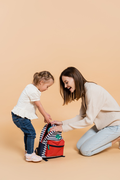 girl with down syndrome and excited mother looking at backpack on beige  - Photo, image