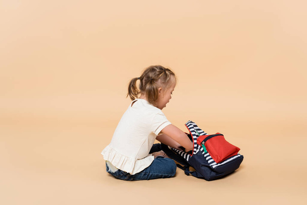 дитина з синдромом Дауна сидить з рюкзаком на бежевому
 - Фото, зображення