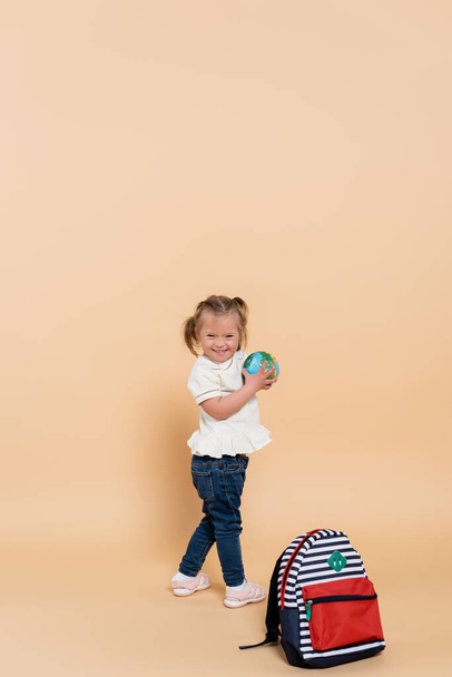 happy kid with down syndrome holding small globe near backpack on beige - 写真・画像