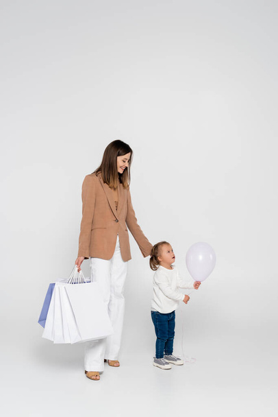 feliz madre sosteniendo bolsas de compras y mirando a la chica con síndrome de Down cerca de globo en gris - Foto, imagen