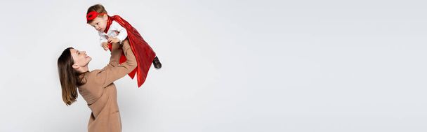 happy mother lifting toddler son in superhero costume isolated on grey, banner - Fotoğraf, Görsel