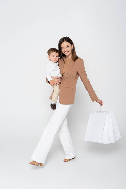 joyful mother holding in arms toddler son and shopping bags while walking on grey - Photo, image