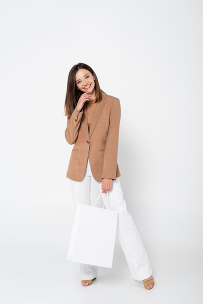 full length of happy young woman in beige blazer holding shopping bag on white - Фото, зображення