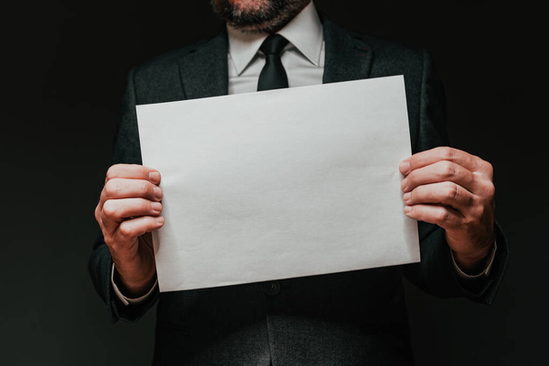 Businessman holding blank white paper as mockup copy space, elegant handsome entrepreneur in dark interior, selective focus - Фото, изображение