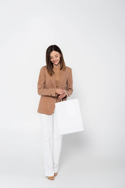 cheerful young woman in beige blazer holding shopping bag while posing on white - Foto, Bild