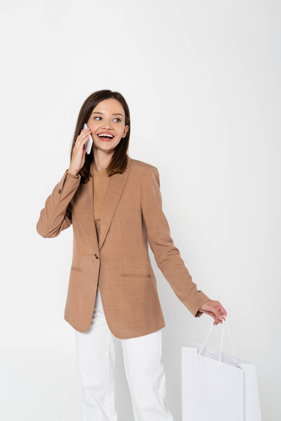 happy young woman in beige blazer holding shopping bag and taking on smartphone on grey - Foto, Imagem
