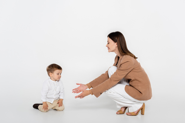 happy woman in beige blazer sitting and reaching toddler boy on grey - Photo, Image
