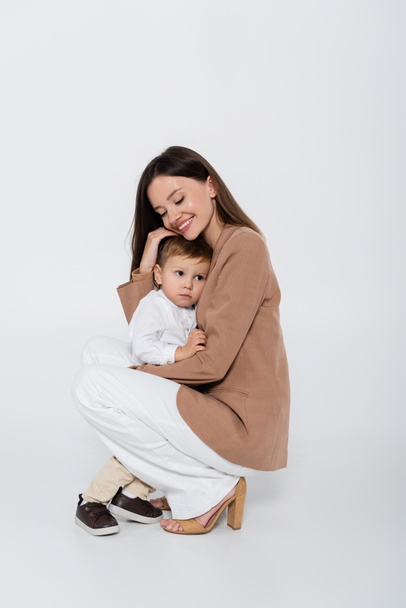 happy woman in beige blazer sitting and hugging toddler boy on grey - Photo, image