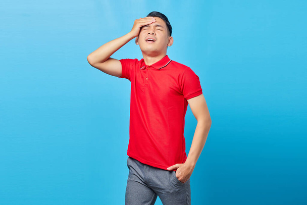 Portrait of adult Asian man in red shirt getting painful migraine gesture and holding head isolated on blue background - Photo, Image