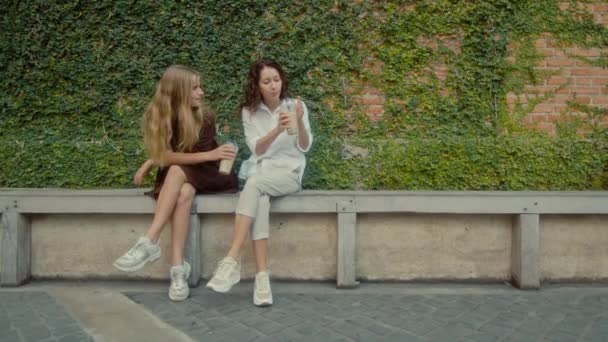 Two women sit on a bench near a wall of vines and drink drinks - Video