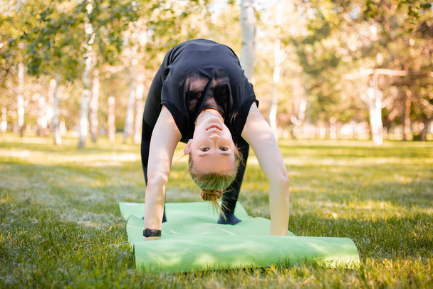 The girl is engaged in yoga, stands in a bridge pose on a sports mat in the park. - Photo, image
