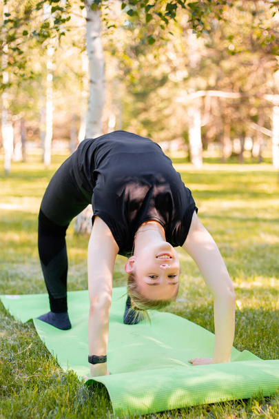 The girl is engaged in yoga, stands in a bridge pose on a sports mat in the park. - Foto, immagini