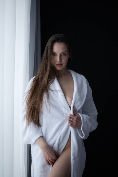 Low angle side view of sexy female with bare breast standing near window in room and looking at camera - Fotografie, Obrázek