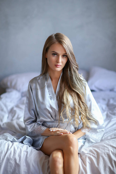 Attractive and sexy young blonde woman in silver nightgown sitting on the bed - Фото, изображение