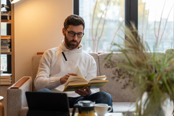 Full length view of the caucasian bearded man wearing glasses sitting at the lonely room during the evening and reading interesting book with calm face - Foto, Imagem