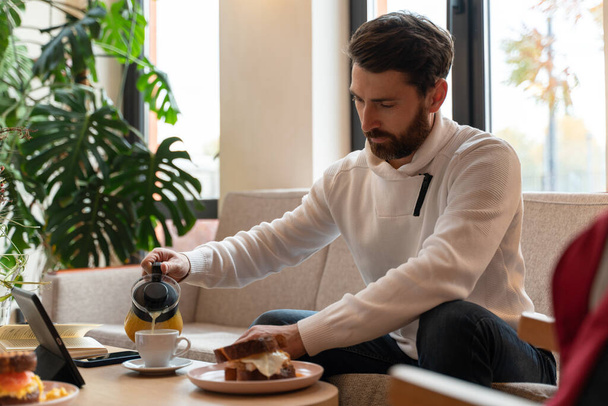 Retrato de un joven guapo sentado en la mesa con la tableta y el trabajo. Él está mirando atentamente mientras vierte café a la taza  - Foto, Imagen