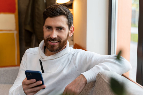 Portrait view of the caucasian man looking at the camera with smartphone at his hands while sitting at the sofa and working or chatting with friends  - Φωτογραφία, εικόνα