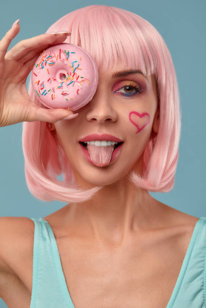 Retrato de una chica de moda con una peluca rosa y con una dona rosa en sus manos mantiene a la vista, muestra la lengua - Foto, imagen