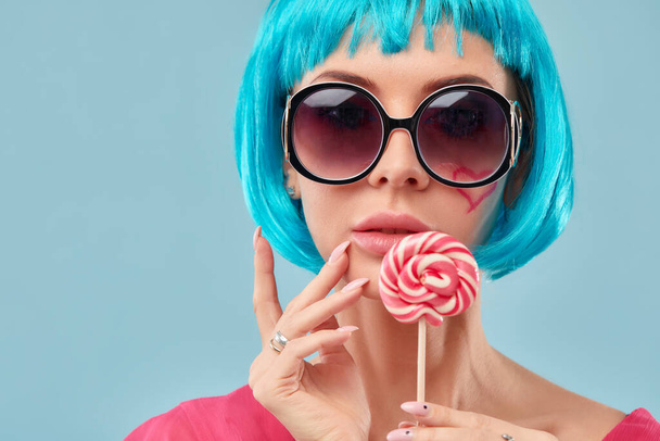 Young funky girl in blue wig and eyeglasses, holding lollipop candy, looking aside with dreamful smile as if making plans for party, isolated on blue background - Φωτογραφία, εικόνα