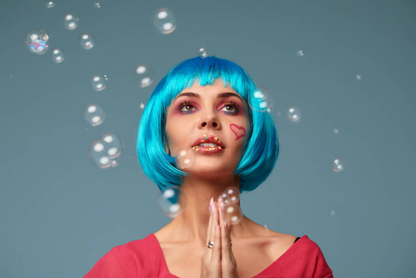 Beautiful young woman with blue wig and bright make-up in soap bubbles hands holds at the chin makes a wish. Fashion model girl with creative color makeup. Women doll concept. - Foto, afbeelding