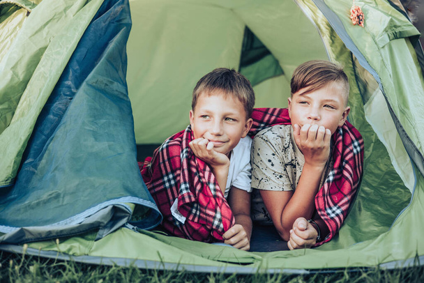 Onnelliset lapset makaamassa ruudullinen teltassa. Perheviikonloppu luonto ulkona. Hymyileviä teinipoikia muki kädessään. Hidas elämä - Valokuva, kuva