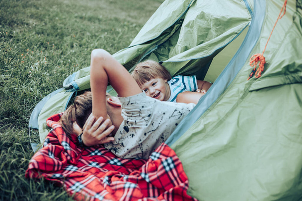 Happy children lying on plaid in camping tent. Family weekend nature outdoor. Smiling teenage boy and toddler having fun. Slow life - Zdjęcie, obraz