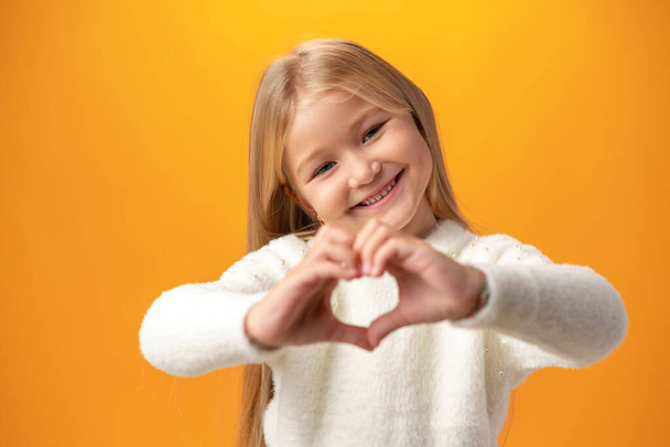 Happy little girl showing heart love gesture over yellow background - Foto, afbeelding