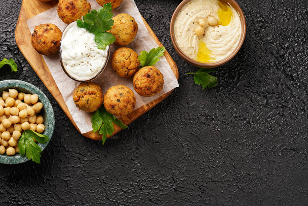 Traditional oriental chickpea deep fried falafel on a wooden board, tzatziki yoghurt sauce, hummus, fresh lime and green cilantro on black surface, copy space - Photo, Image