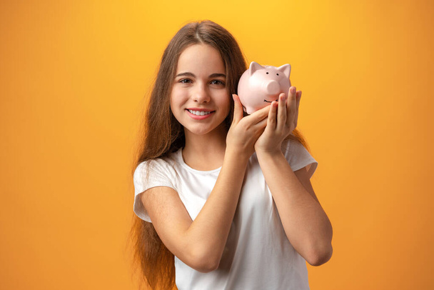 Teen girl with piggy bank on yellow background. - Fotoğraf, Görsel