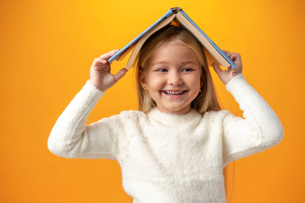 Little girl holding book on her head and looking at camera over yellow background - Foto, afbeelding