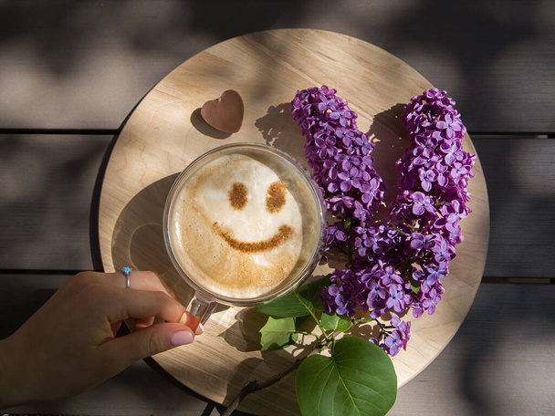 Lilac, a female hand near a cup of coffee with a smile from cinnamon on milk foam on a round wooden stand. View from above. Positive thinking, good morning, hello spring - Fotoğraf, Görsel