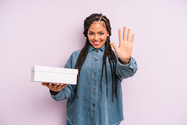negro afro mujer sonriendo y buscando amigable, mostrando el número cinco. concepto de caja de paquetes - Foto, Imagen