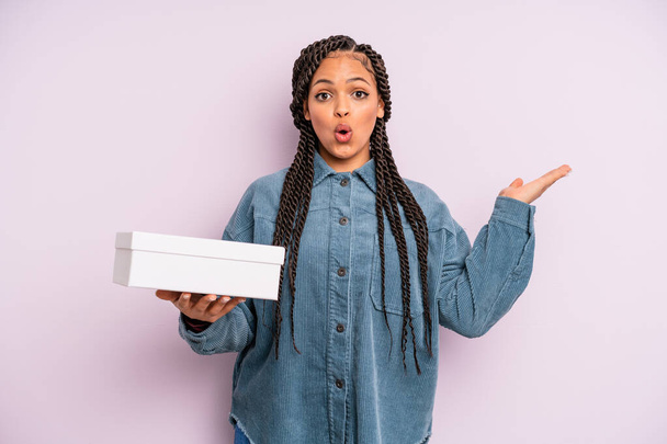 black afro woman looking surprised and shocked, with jaw dropped holding an object. packages box concept - Fotoğraf, Görsel