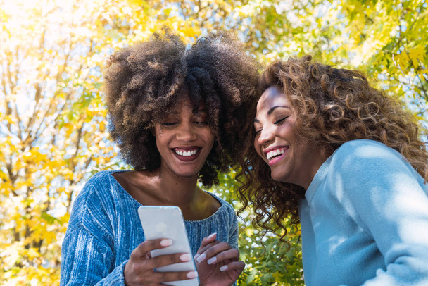 Дві жінки з афроамериканських друзів перевіряють, як соціальні мережі тримають смартфон на відкритому повітрі. Чорні друзі з кучерявим волоссям і синім джемпером посміхаються під час використання мобільного телефону і розважаються в парку
 - Фото, зображення