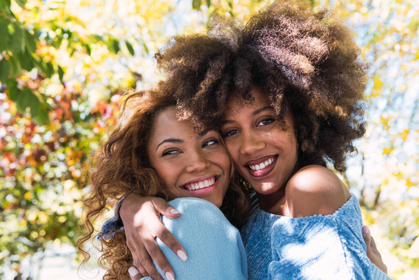 İki Afro-Amerikalı kadının yakın çekimi ve mutlu en iyi arkadaşlar birbirlerine sarılıp parkta gülüyorlar. Kıvırcık saçlı, sevgi dolu gülümseyen ve sıcacık sarılan neşeli afro siyahi arkadaşlar. - Fotoğraf, Görsel