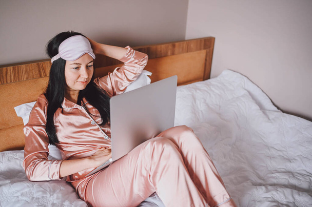 Young happy brunette woman in sleeping mask lying in bed in pink silk pajamas with laptop computer, have online video chat. Blogger or freelancer with remote work at home. - Photo, Image