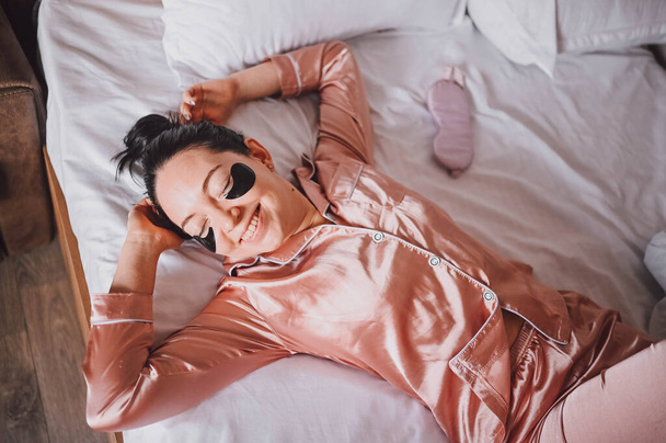 Cosmétologie, soin de la peau, soin du visage, spa, concept de beauté naturelle. Une jeune femme souriante en pyjama de soie rose se trouve au lit avec des taches hydratantes noires sur les yeux. routine beauté. - Photo, image