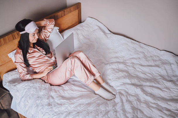 Νεαρή ευτυχισμένη μελαχρινή γυναίκα με μάσκα ύπνου ξαπλωμένη στο κρεβάτι με ροζ μεταξωτές πιτζάμες με φορητό υπολογιστή, έχουν online video chat. Blogger ή ελεύθερος επαγγελματίας με απομακρυσμένη εργασία στο σπίτι. - Φωτογραφία, εικόνα