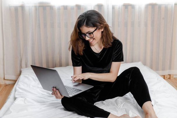 Good morning concept. Beautiful sexy young brunette girl with glasses wearing a black velvet pajama sleepwear on white bed in light cozy bedroom with laptop - Foto, Imagen