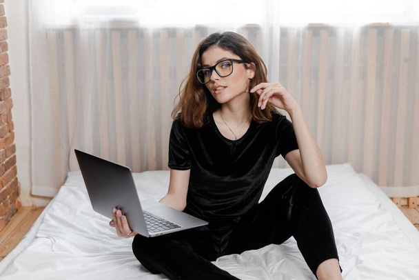Καλημέρα έννοια. Όμορφη σέξι νεαρή μελαχρινή κοπέλα με γυαλιά φορώντας ένα μαύρο βελούδινο πιτζάμα σλιπ σε λευκό κρεβάτι στο αναπαυτικό υπνοδωμάτιο με φορητό υπολογιστή - Φωτογραφία, εικόνα