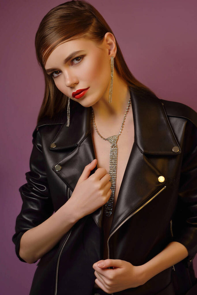 A young beautiful girl posing in a black leather jacket and gold jewelry on a background in a photo studio. - Foto, Imagem