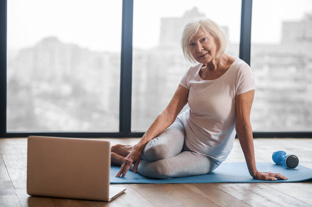Gray-haired senior woman sitting on the floor and having an online yoga class - Zdjęcie, obraz