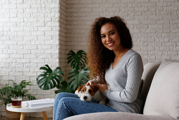 Portrait of young beautiful black woman with her adorable wire haired Jack Russel terrier puppy at home. Loving girl with rough coated pup having fun on the couch. Background, close up, copy space. - Fotoğraf, Görsel