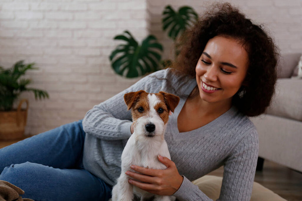 Portrait of young beautiful black woman with her adorable wire haired Jack Russel terrier puppy at home. Loving girl with rough coated pup having fun on the couch. Background, close up, copy space. - Foto, afbeelding