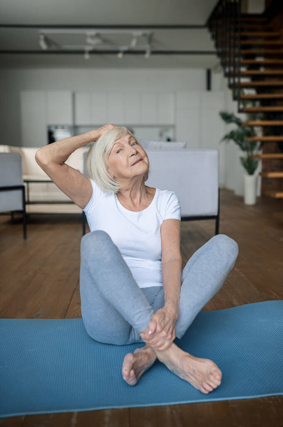 Старша жінка сидить на килимку і виконує вправи
 - Фото, зображення