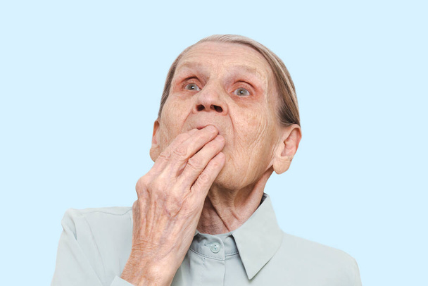 Lifestyle portrait of an elderly woman with a hand covering her mouth on a blue background. Surprised old lady pensioner with a frightened expression on her face. Concept of emotional shock. - 写真・画像