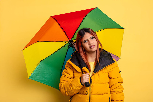 hispanic woman feeling puzzled and confused. rain and umbrella concept - Foto, Imagen