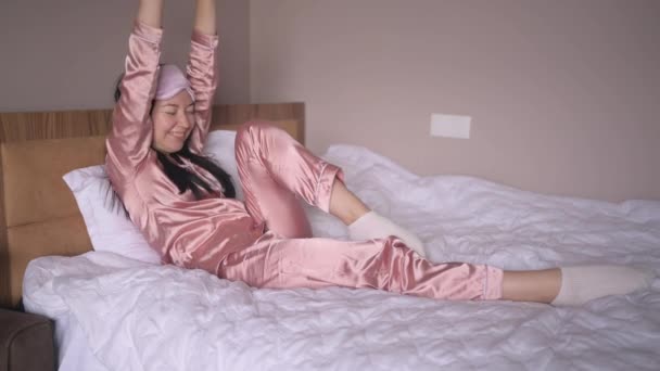 Atrakcyjna uśmiechnięta młoda kobieta w różowej jedwabnej piżamie i masce do spania oczy rozciągająca się w łóżku budząca się sama szczęśliwa koncepcja, obudzona po zdrowym śnie w wygodnym łóżku i materacu cieszyć się porankiem. - Materiał filmowy, wideo