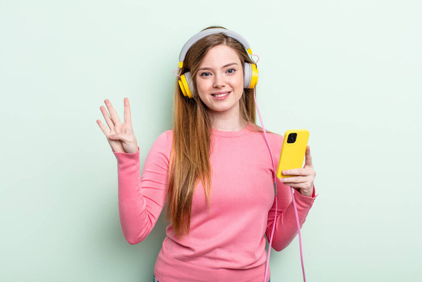 redhair woman smiling and looking friendly, showing number four. headphones and smartphone concept - Photo, Image