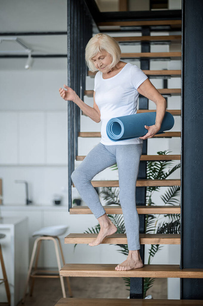 A senior woman getting downstairs with a yoga mat in hands - Zdjęcie, obraz